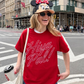 Person wearing a red 'Hiya Pal!' t-shirt and Minnie Mouse hat on a city street.