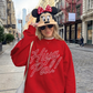Person wearing a red sweatshirt with 'Hiya Pal!' text, Minnie Mouse hat, and sunglasses on a city street.