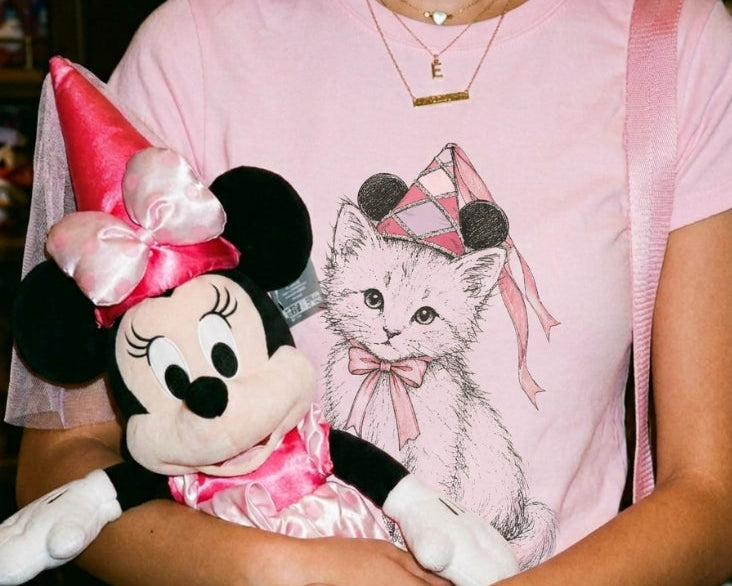 A person wearing a light pink graphic T-shirt with a Minnie Mouse and kitten print, holding a Minnie Mouse plush toy. The background is a blurred indoor retail setting.