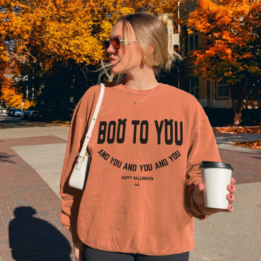 A woman stands outdoors on a sidewalk in front of autumn trees, wearing a terracotta-colored sweatshirt with black text that reads 'BOO TO YOU AND YOU AND YOU AND YOU' and 'HAPPY HALLOWEEN' below it. She is also wearing black shorts, holding a white coffee cup, and carrying a white shoulder bag.
