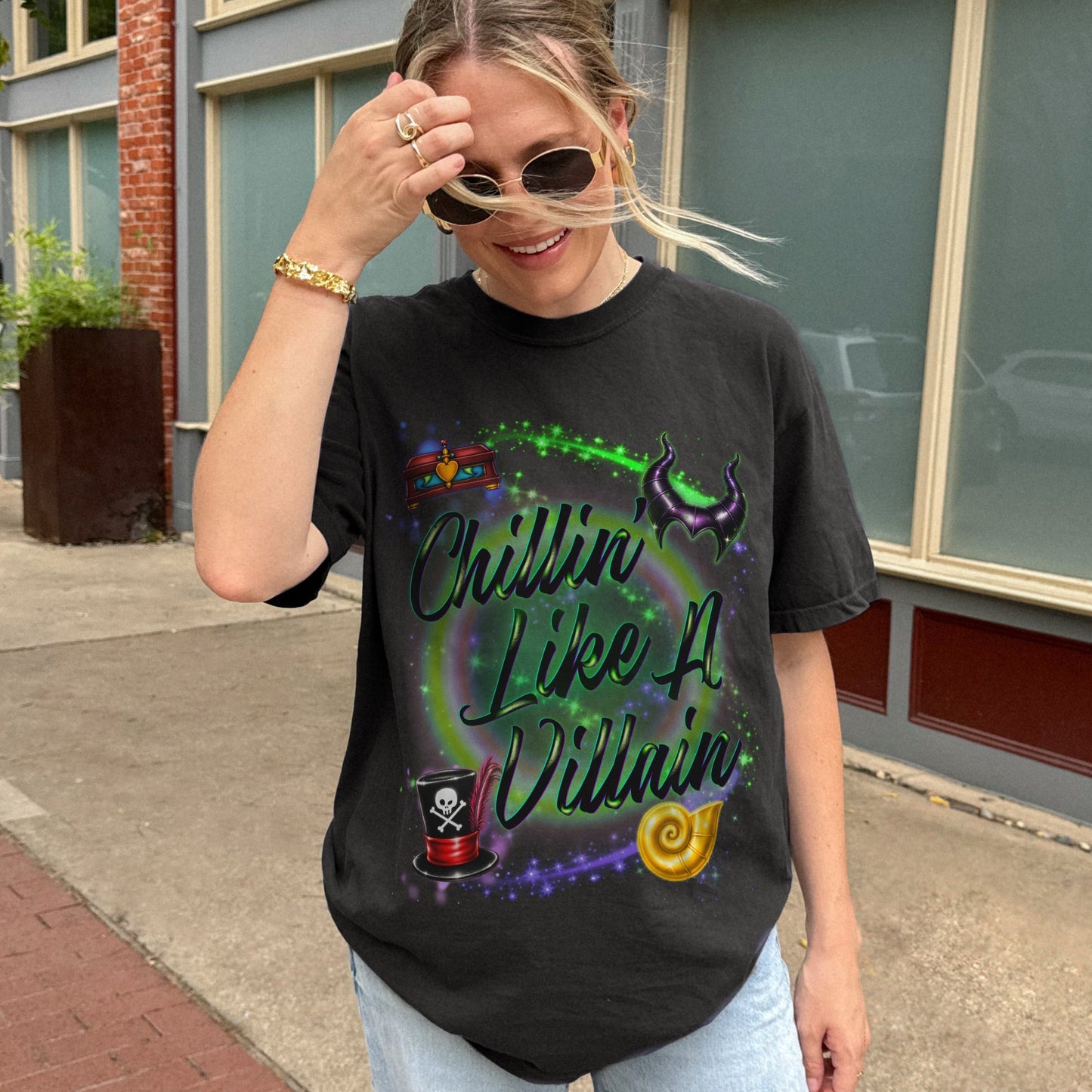 A woman wearing a black graphic t-shirt with a colorful retro-style design that reads 'Chillin' Like A Villain' and light wash blue jeans, standing outdoors on a sidewalk in front of a brick and gray building.