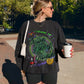 A woman wearing a black graphic sweatshirt with 'Chillin Like A Villain' in green script, standing outdoors on a brick sidewalk holding a white coffee cup.