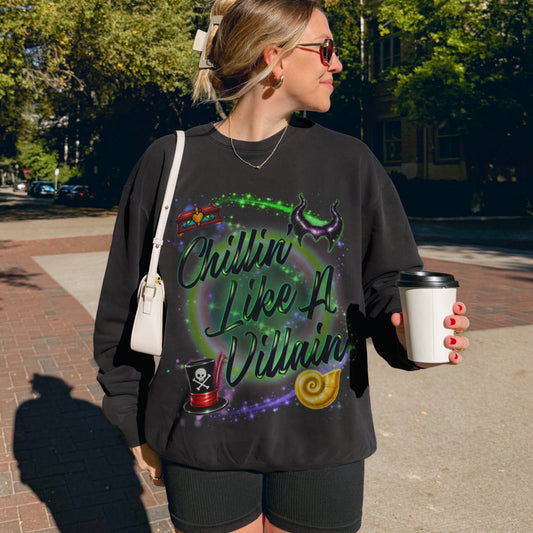 A woman wearing a black graphic sweatshirt with 'Chillin Like A Villain' in green script, standing outdoors on a brick sidewalk holding a white coffee cup.