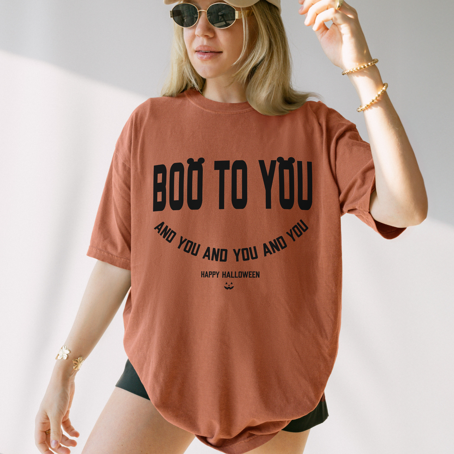 A woman modeling a terracotta-colored graphic t-shirt with 'BOO TO YOU' printed in black. She is wearing a beige baseball cap, round sunglasses, and gold jewelry against a bright, sunlit white background.