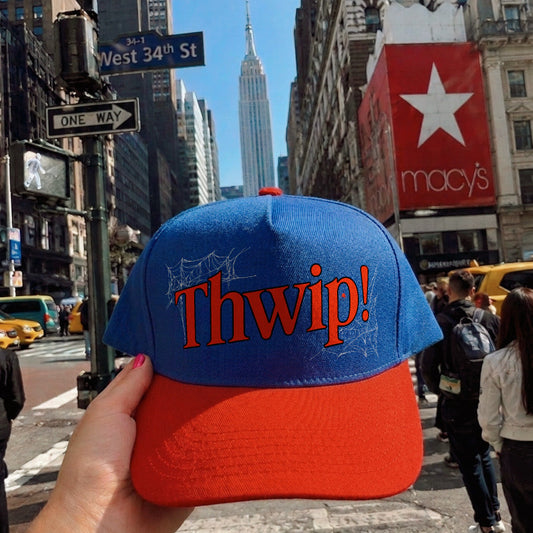 A blue and red baseball cap with 'Thwip!' embroidered on the front, held by a hand in a busy New York City street scene with pedestrians and buildings in the background.